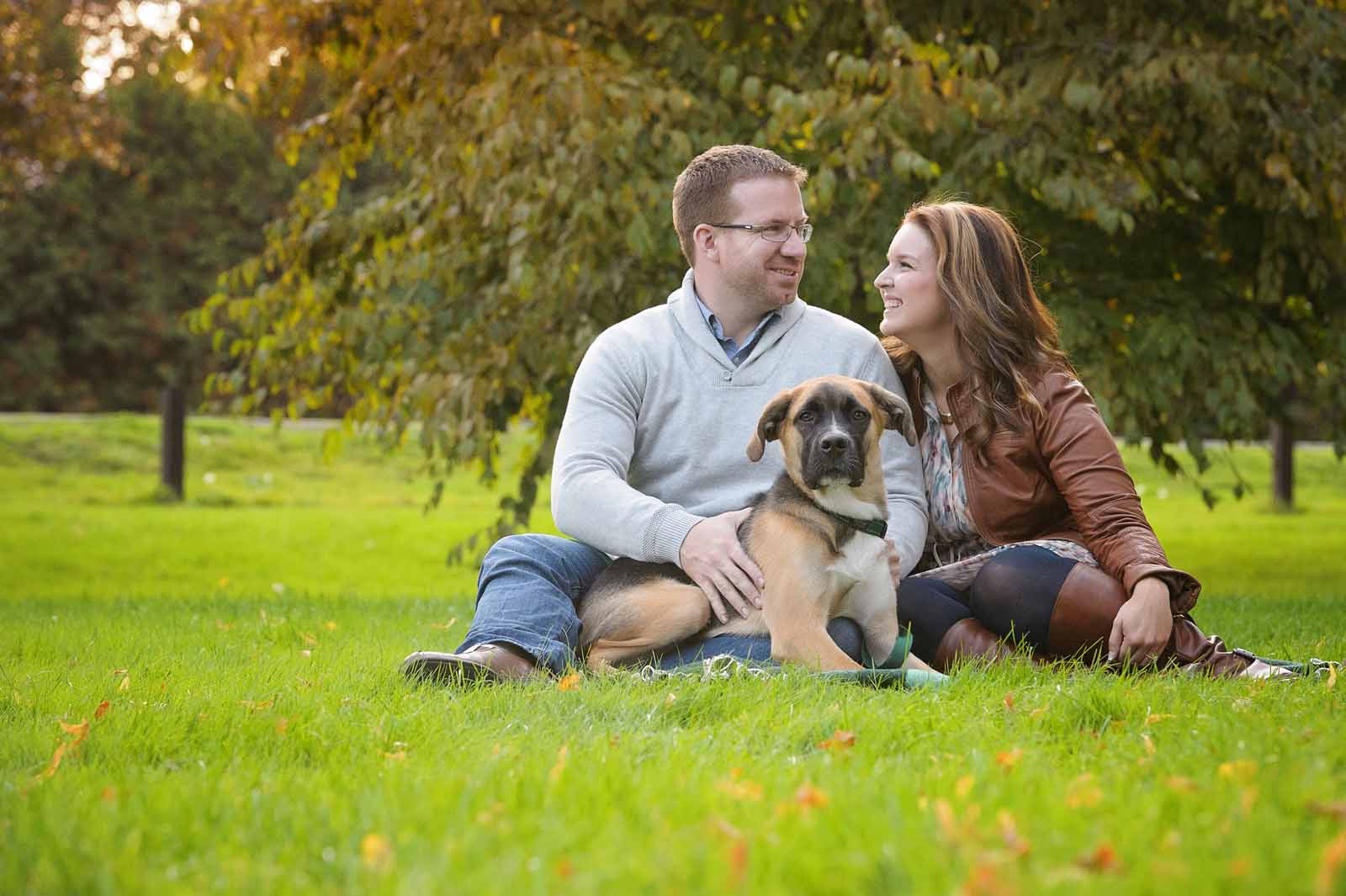 An engaged couple sitting on the grass with their dog, a lab mix, looking at each other with love at the Arboretum in Ottawa