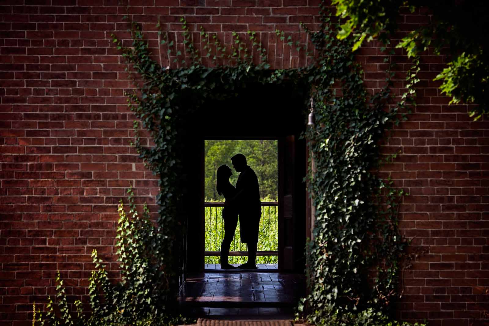 An engaged couple standing looking at each other, silhouetted against the vineyard background behind them, surrounded by the brick facade of the winery where their engagement session took place