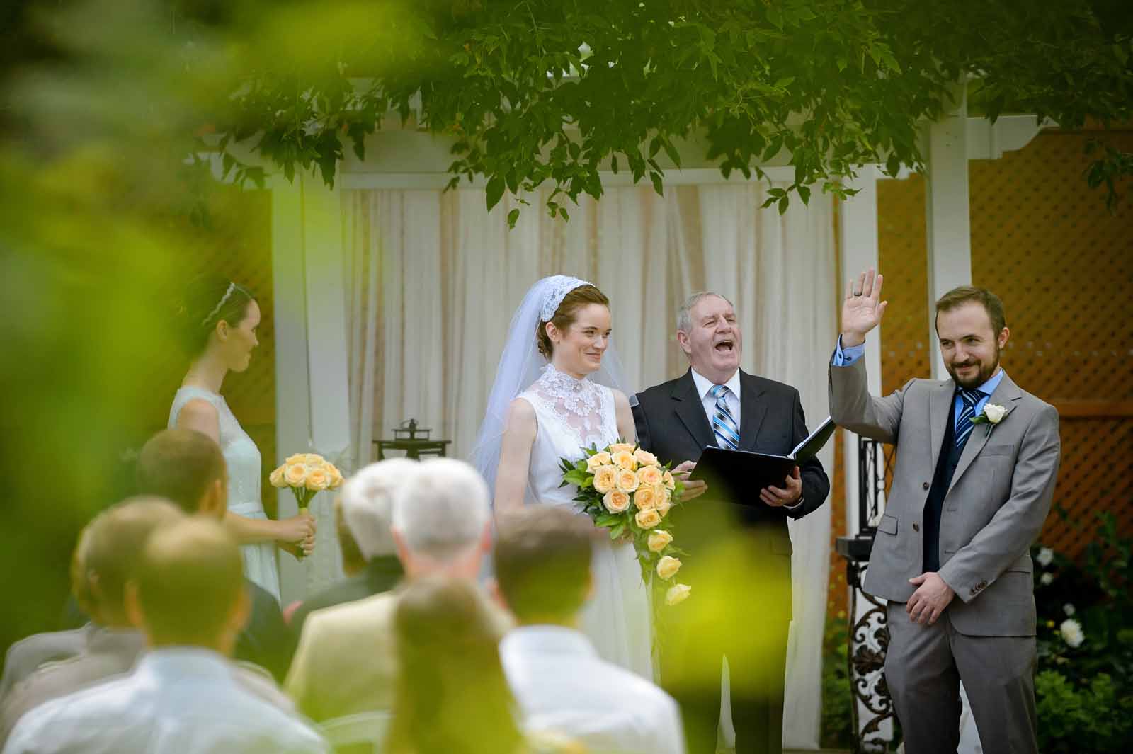 A groom cracking a joke, while his new bride smirks at him, at the officiant laughs loudly during an outdoor ceremony at The Schoolhouse in Munster, Ottawa, Ontario