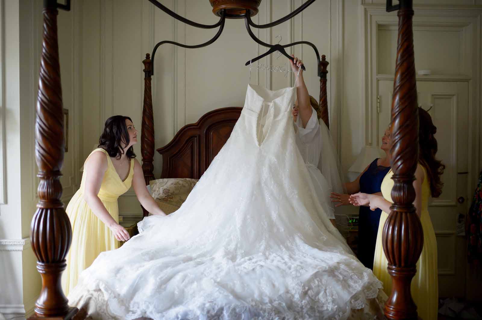 Wedding dress hanging on a bed and being perfected by bridesmaids while getting ready for the ceremony