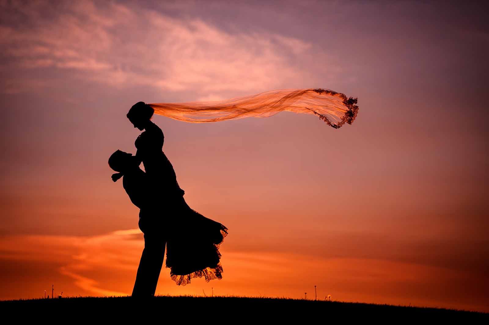 Silhouette with a pink sky of the groom lifting the bride into the air and her veil floating behind her